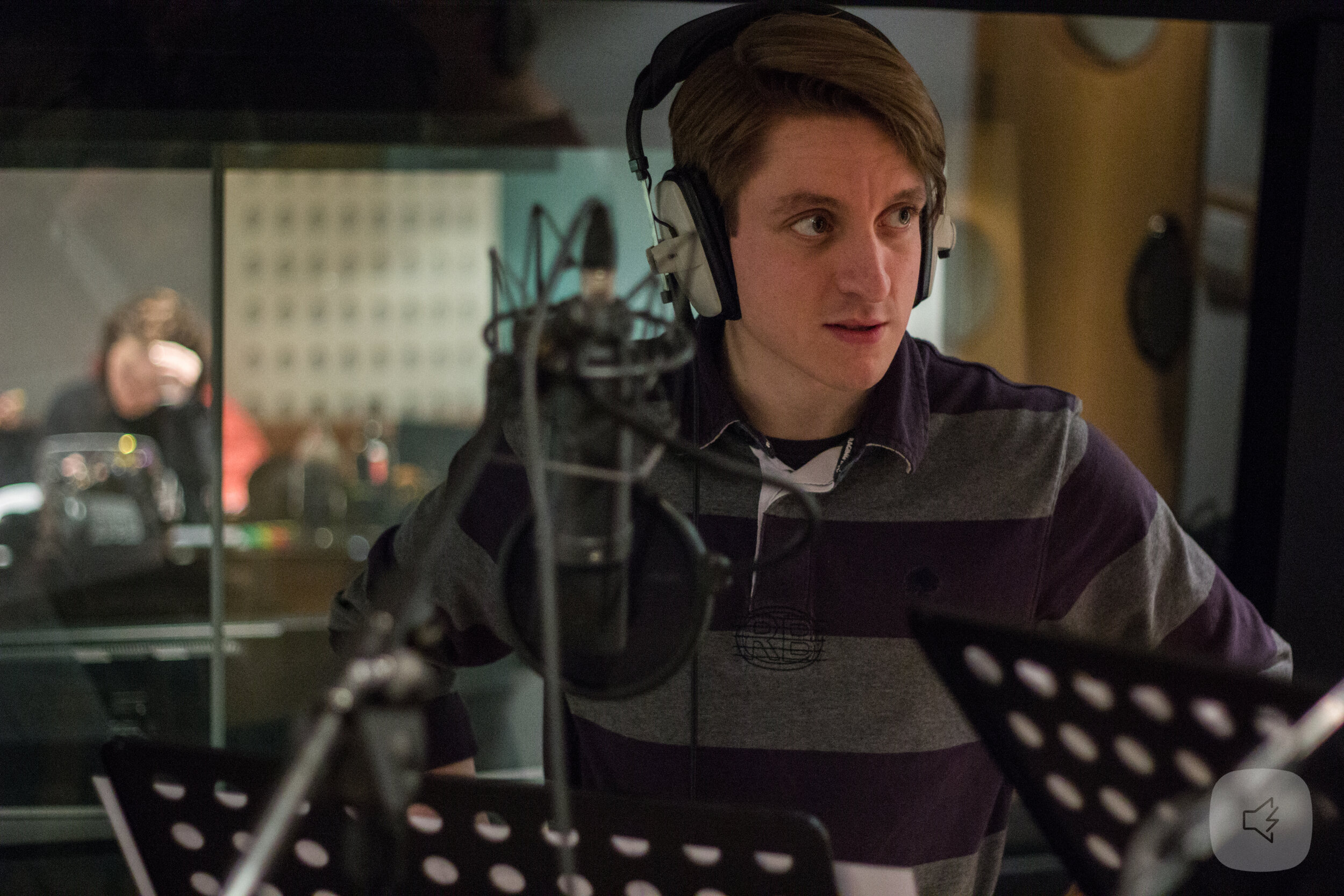 colour photo. taken inside a sound recording room. matthew is in front of a microphone and is wearing headphones. he is wearing a shirt that is striped horizontally with grey and dark purple.