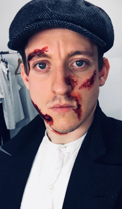 colour photo. matthew looks into the camera, dressed as banquo in a black jacket and white shirt with a navy blue flat cap on his head. his face is covered with realistic makeup of many bloody stab wounds.