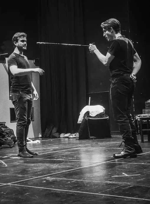 black and white photo. two actors stand facing each other in a mildly cluttered rehearsal space. matthew stands on the right, pointing at the other actor with a cane.