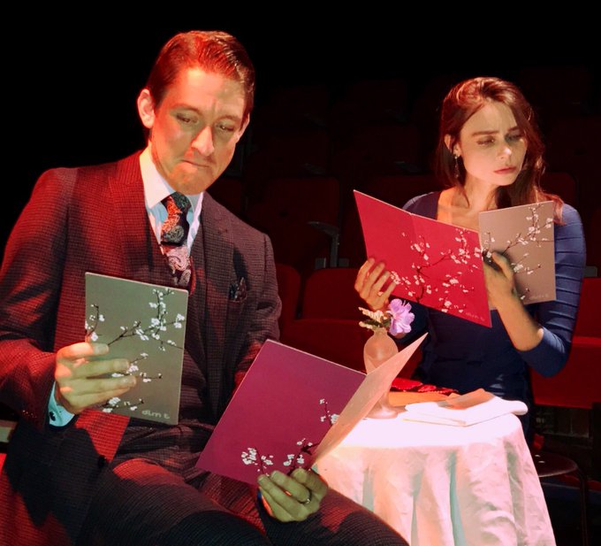 colour photo. matthew's character and another, female character are seated at a table in formal wear. both of them consider menus with cherry blossom designs on. the woman looks soulfully contemplative, while matthew's character smiles slightly.
