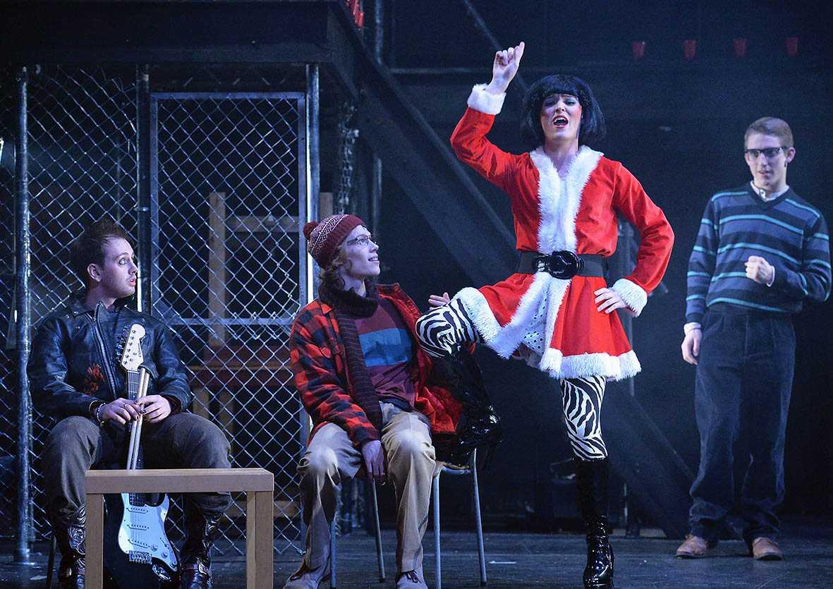 colour photo. four characters on a grey, industrial set, each dressed very differently. there is a punk man, a man in a cardigan and a beanie who wears glasses and a man in drag as a woman in a santa-esque outfit. tom collins stands on the far right in a jumper and glasses. all actors are looking at the man in drag.
